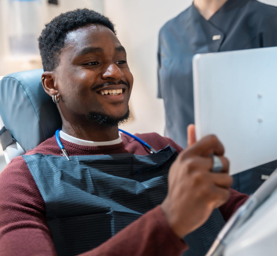 A young black man holding a mirror, looking at his teeth.