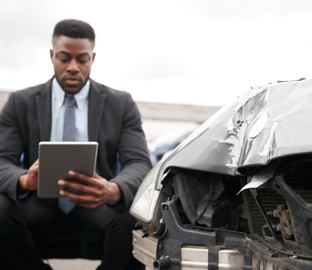 A young black man holding an ipad, taking notes next to a damaged vehicle.