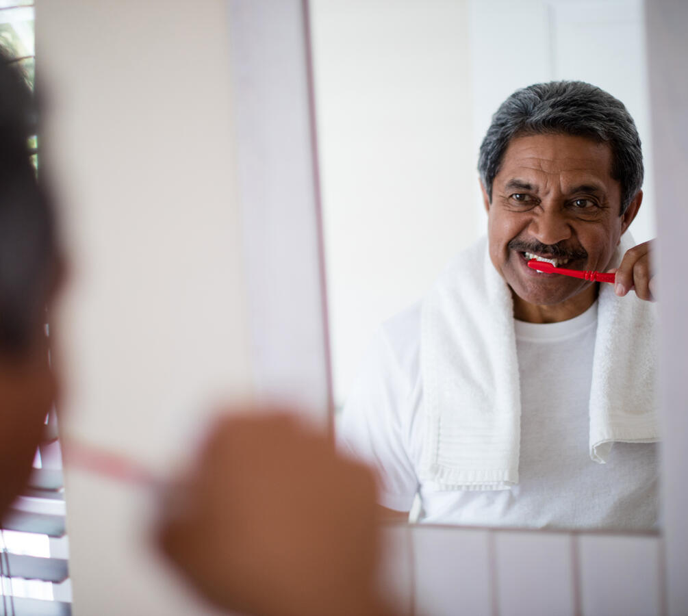 An elderly man brushing his teeth, while looking into a mirror.